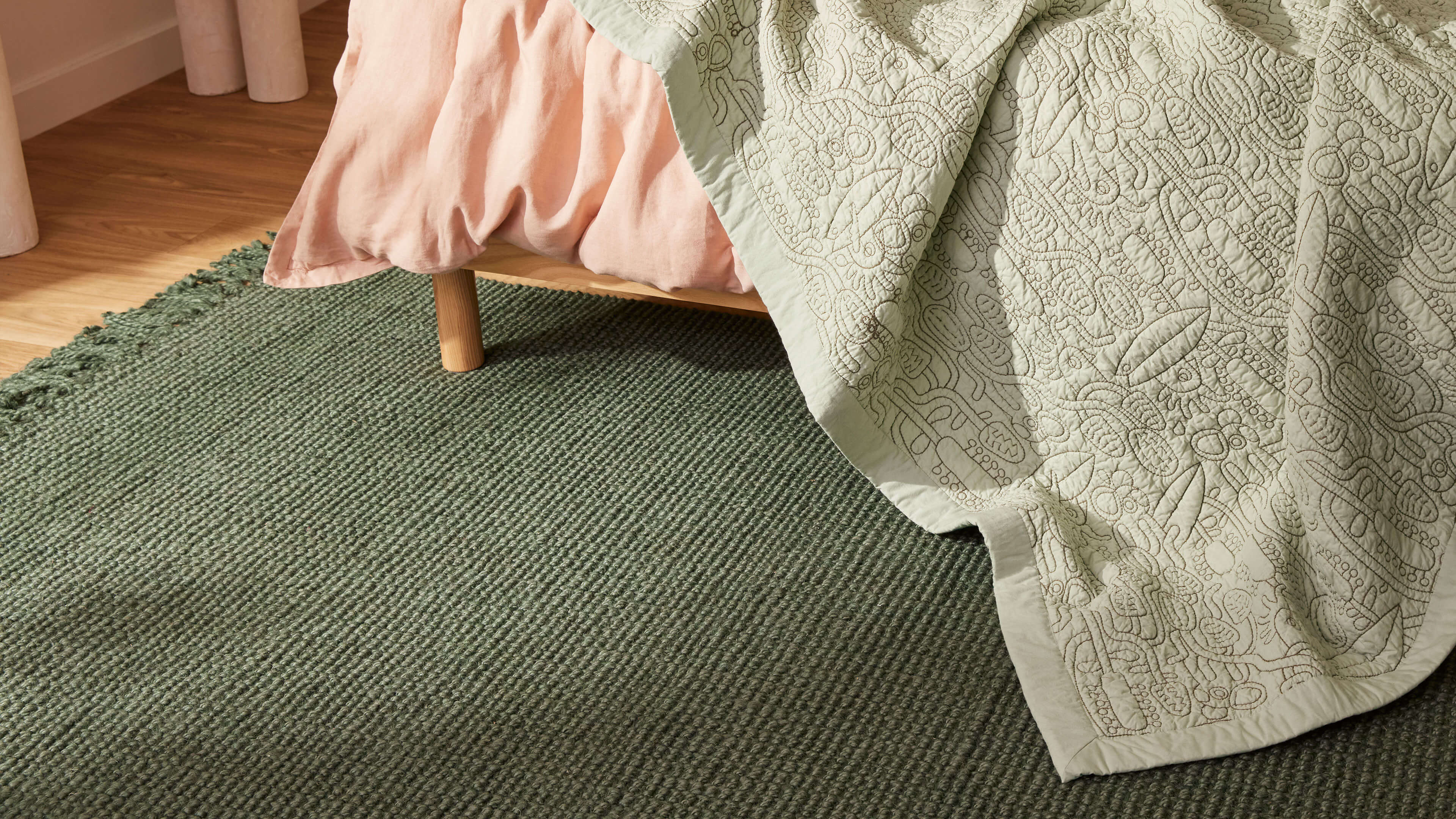 Bedroom scene with a green textured rug, light brown timber floor, and pale green quilt with floral patterns draped over a bed with a pink blanket.