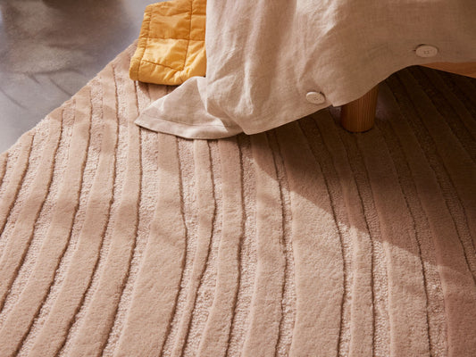 Beige textured rug with striped pattern beneath a light-coloured fabric chair or bed. A yellow quilt is partially visible.
