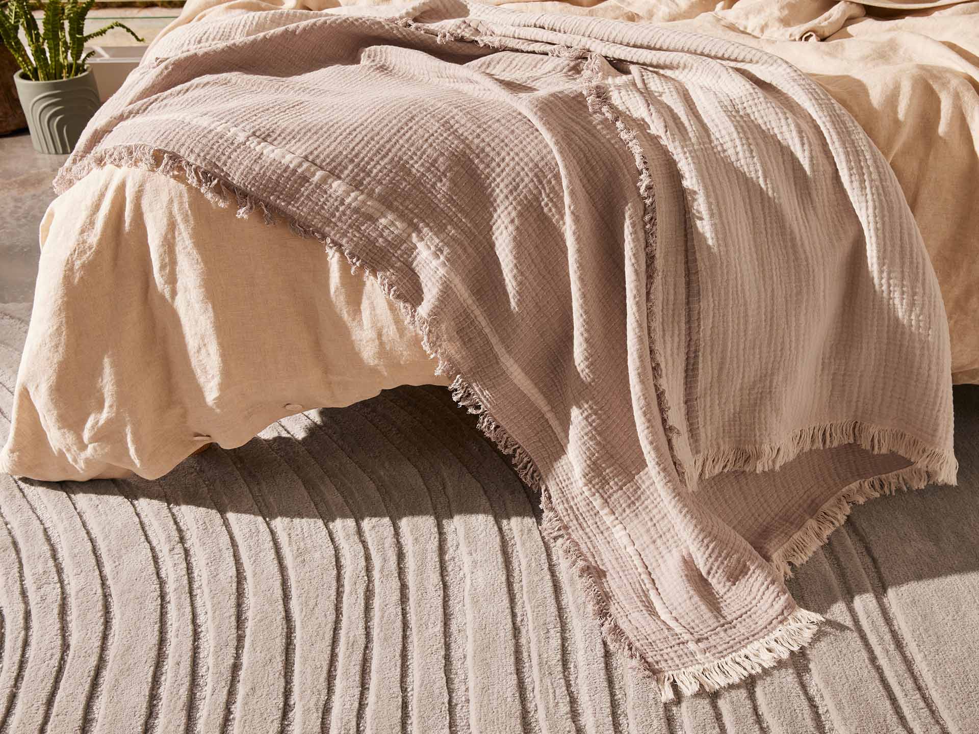 Bed with beige covers and a textured, fringed light taupe throw. Wavy-patterned grey carpet below; plant in the backdrop.