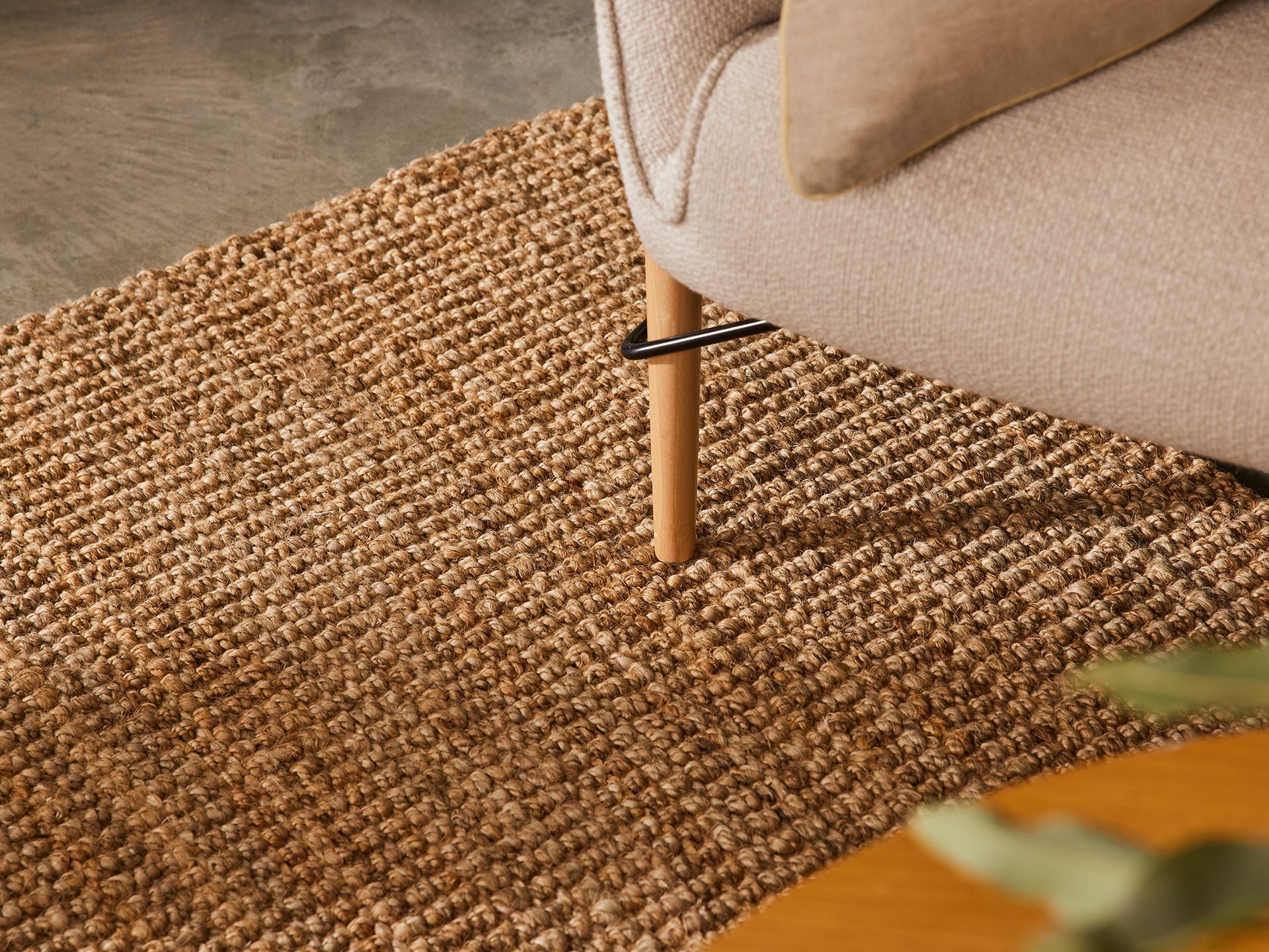 Close-up of a beige sofa leg on a textured brown jute rug, with a glimpse of a light wooden table.