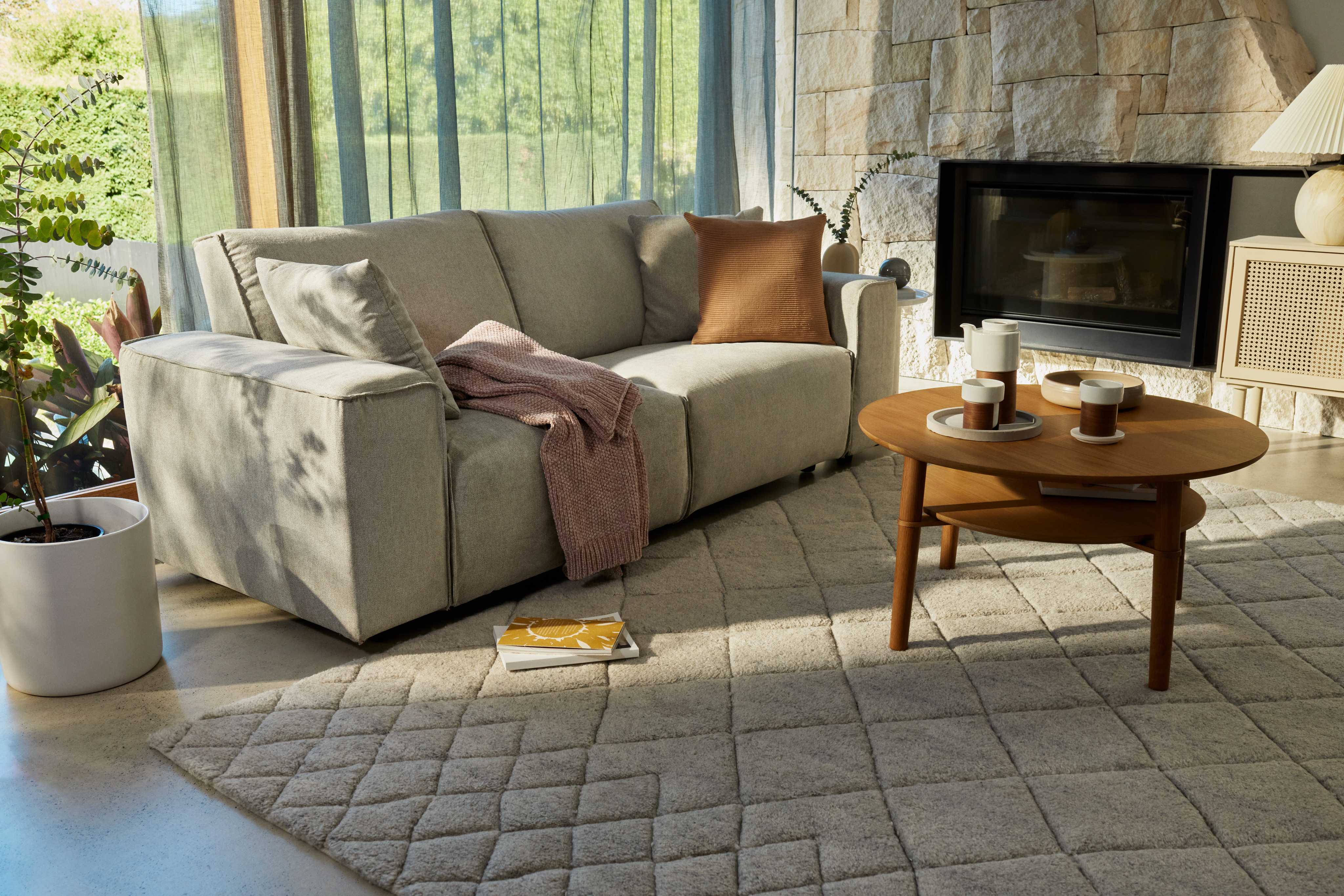 Cosy living room with a light grey couch, brown throw pillow, pink blanket, wooden coffee table, and textured light grey rug, near a fireplace.