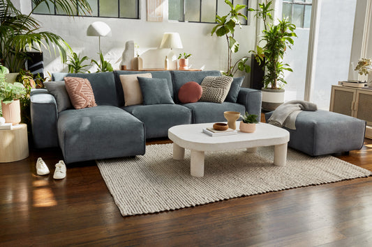 Modern living room with a large grey sectional sofa, assorted cushions, white coffee table on a beige rug, wooden floor, and lush green plants.