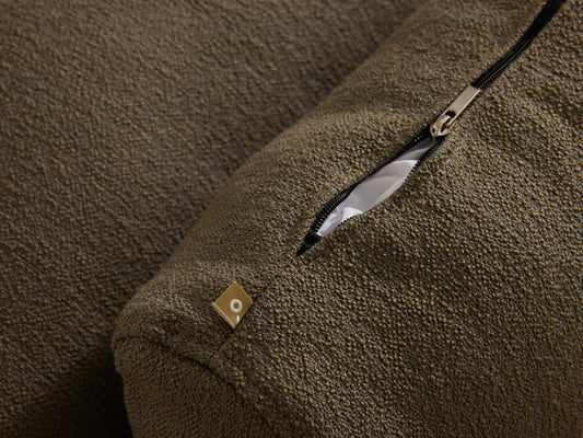 Close-up of a textured dark green cushion with a partially open silver zipper revealing white fabric inside; small decorative tag attached.