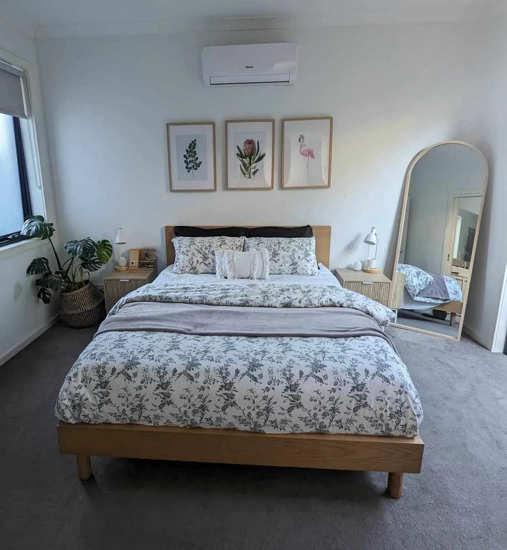 Bedroom with a wooden bed, grey floral bedding, and tall mirror. Wall art above bed and indoor plants beside bedside tables. Neutral tones.