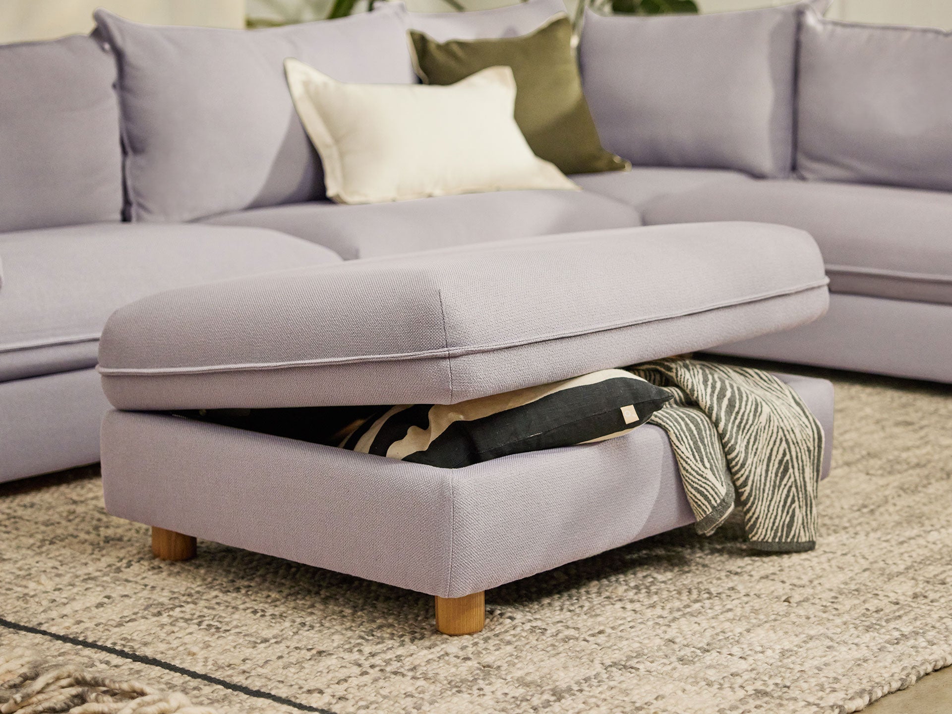 Light grey ottoman with wooden legs used as storage. Grey rug beneath, and matching sectional sofa in the background with decorative cushions.