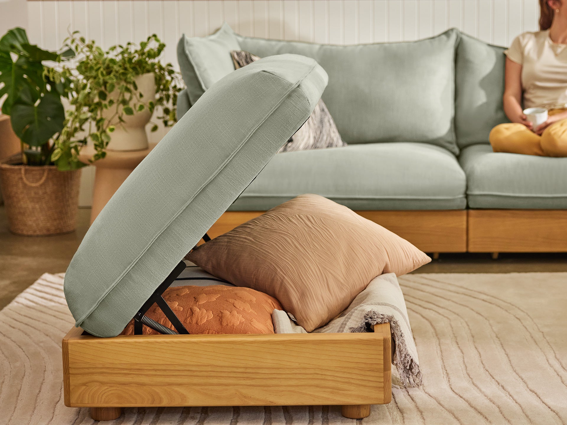 Storage ottoman with a light green cushion top, open to reveal pillows. Light green couch and plants in the background.