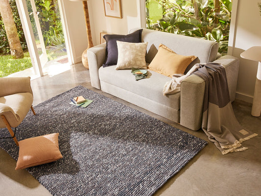 A light grey sofa with assorted cushions, a beige armchair, a dark grey rug, and a draped throw in a bright living room with garden views.