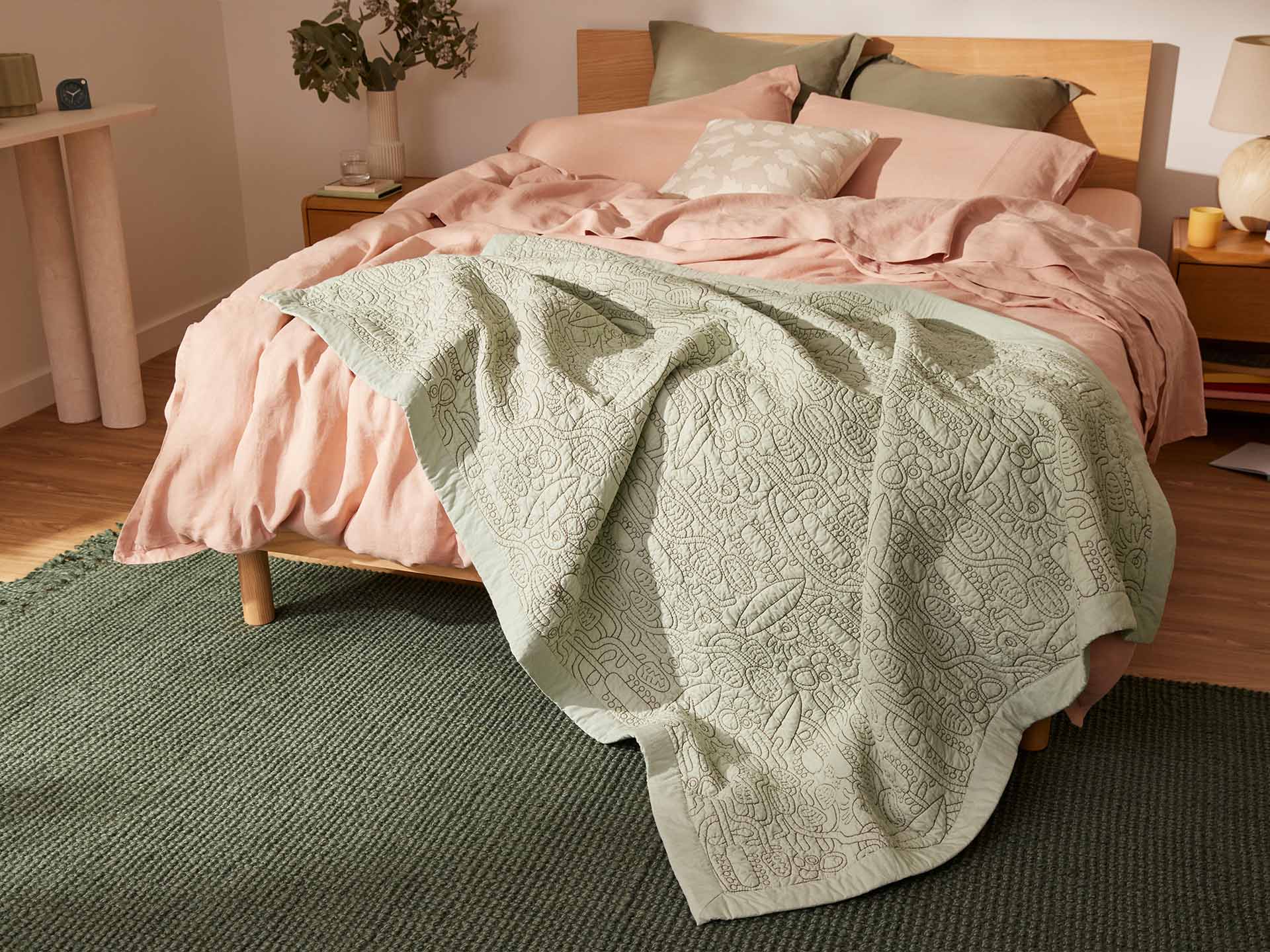 Bedroom scene with a wooden bed, pink bedding, and a light green patterned quilt. A dark green rug lies on a wooden floor.