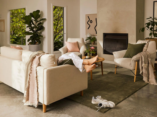 Modern living room with beige sofa, armchairs, green rug, indoor plants, and wall art. Sneakers on floor, natural light from large windows.