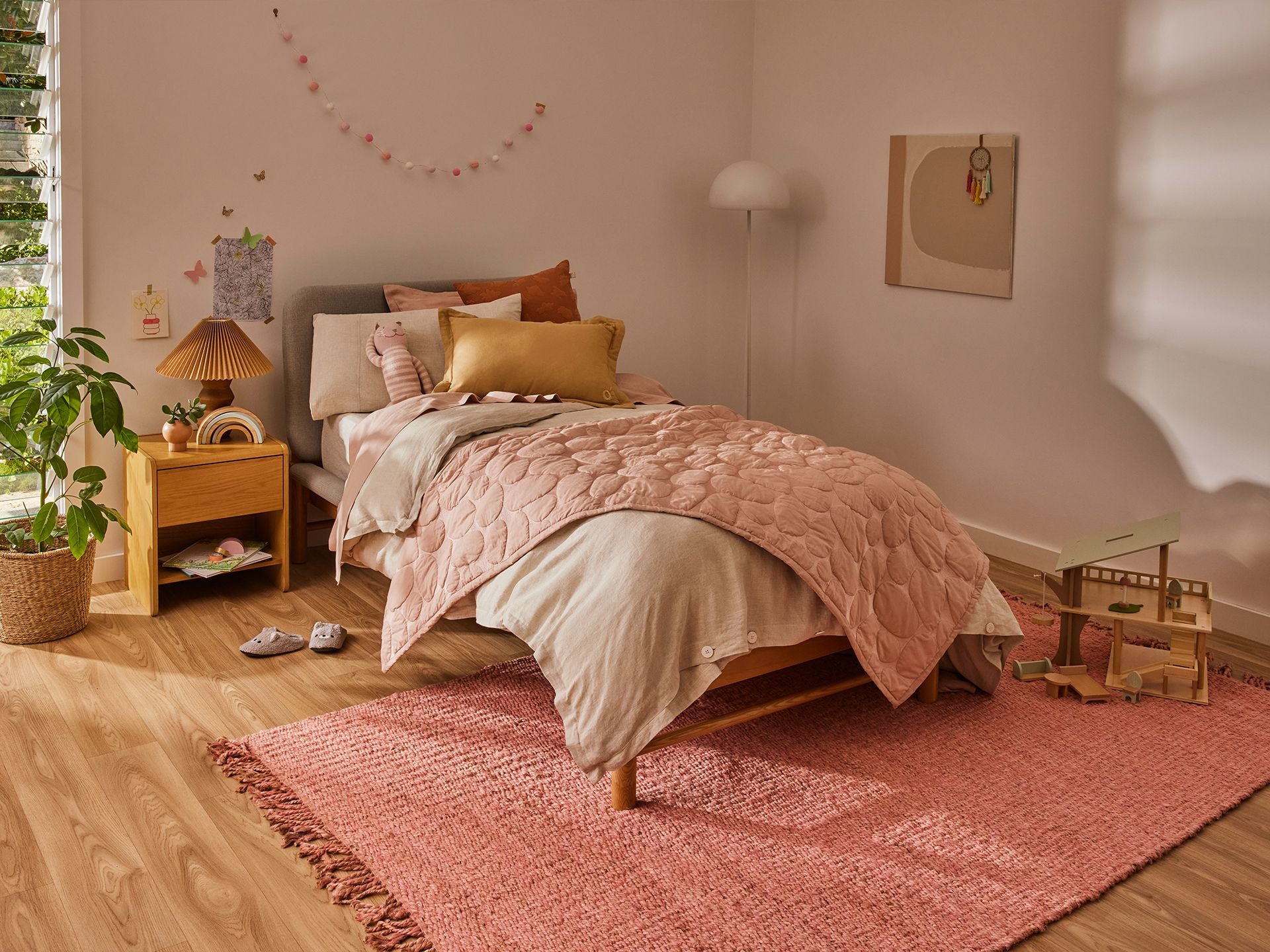 Cosy bedroom with a single bed featuring pink bedding and cushions. A wooden bedside table, lamp, and plant add warmth; a pink rug lies beneath.