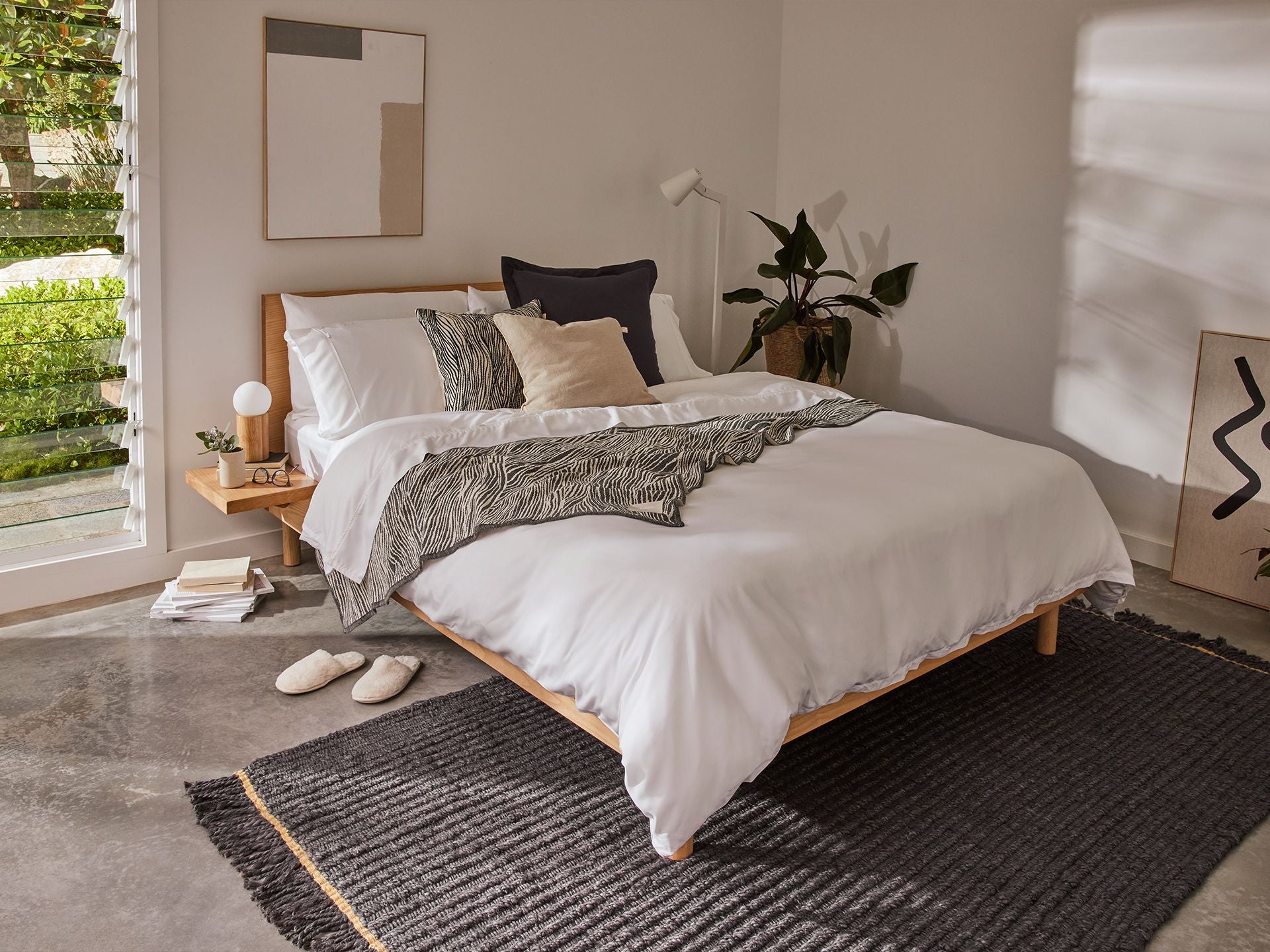 Modern bedroom with a wooden bed frame, white bedding, and grey throw blanket. Black rug on concrete floor, potted plant, and abstract wall art.
