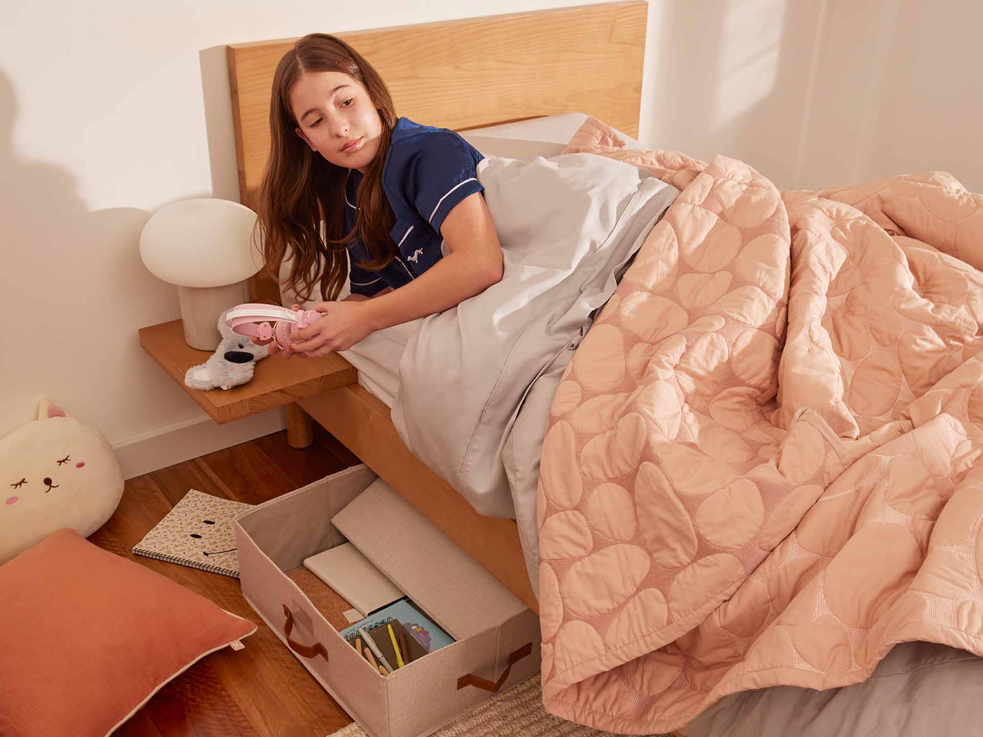 Girl in blue pyjamas on light wooden bed with pink quilt, holding a game controller. Storage box under bed, and cushions on floor.