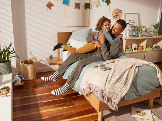 Father and child sitting on a light wood bed with blue and cream bedding, striped socks, indoor plants, and hardwood floor.