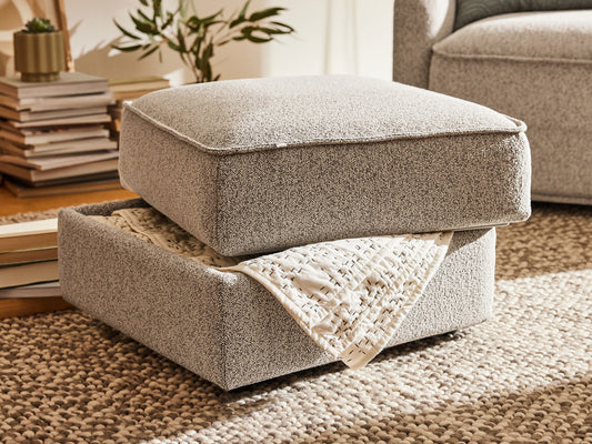Light grey ottoman with textured fabric, showcasing a removable lid and storage, resting on a textured beige rug. Books and a plant in the background.