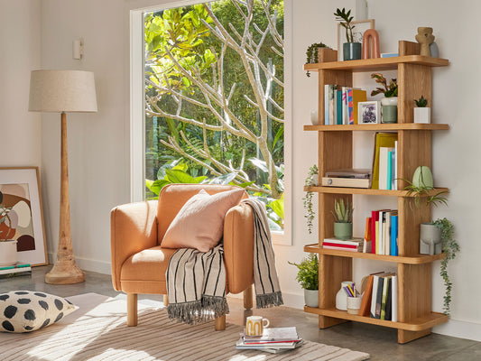 Cosy living room with an orange armchair, striped throw, wooden floor lamp, and bookshelf. Natural light from a large window brightens the space.