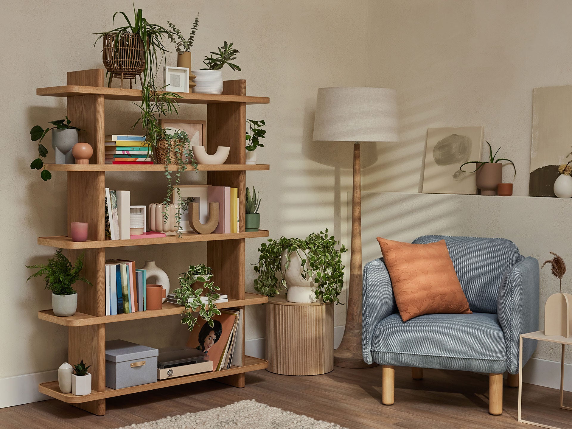 Cosy living room with a blue armchair, orange cushion, wooden bookshelf filled with plants and décor, and a floor lamp. Neutral walls.