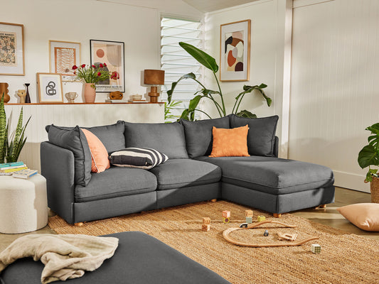 A dark grey L-shaped sectional couch with orange and striped cushions on a beige rug. Decor items, plants, and framed art in the background.