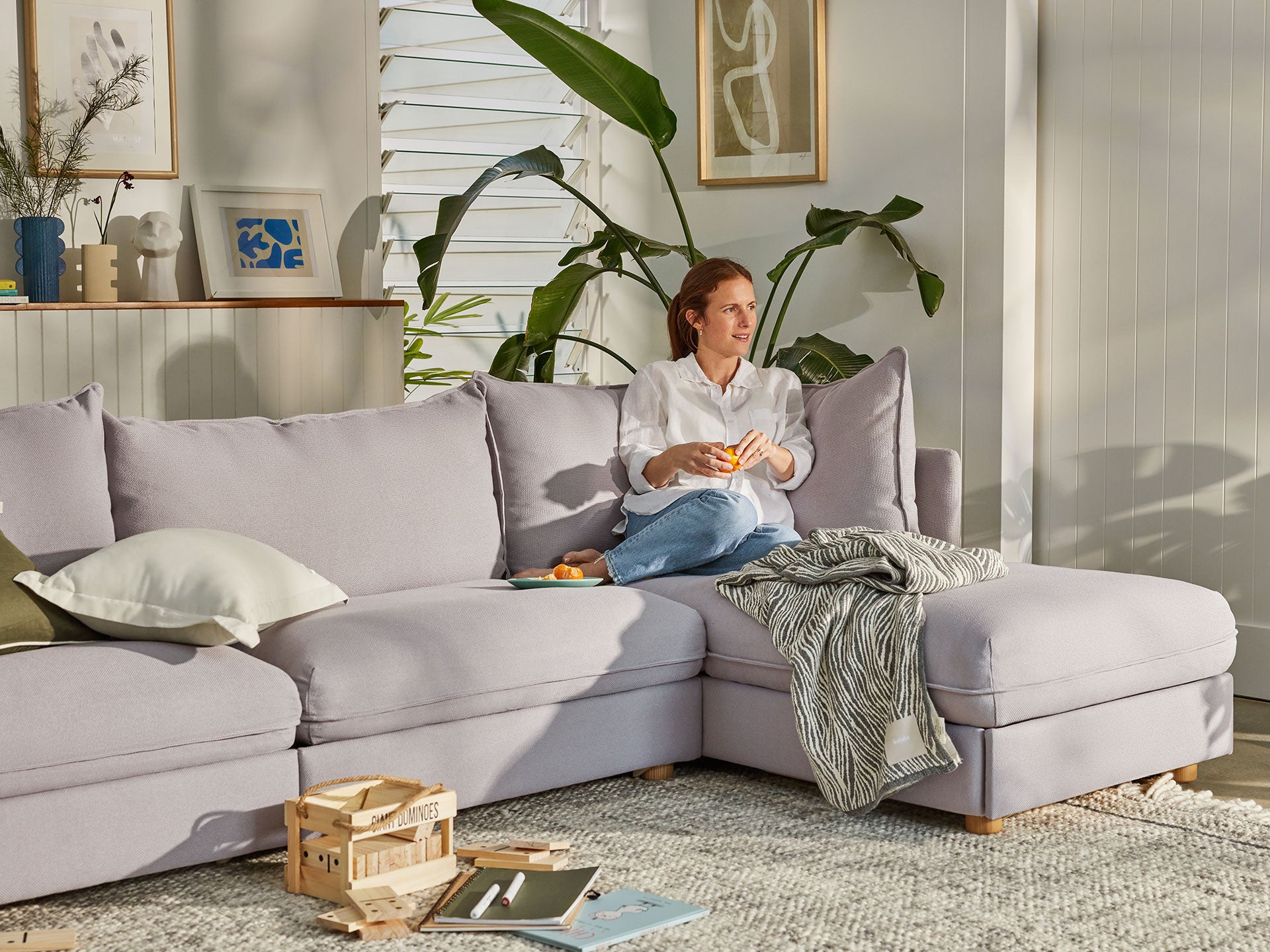 Woman sitting on light grey L-shaped sofa with cushions and a striped throw, surrounded by plants and artwork in a bright living room.