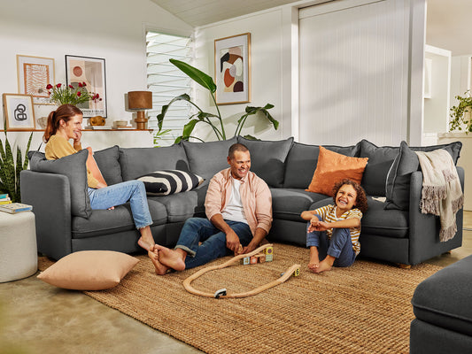 Family sitting on a dark grey sectional sofa with orange cushions, surrounded by artwork and plants, on a jute rug in a cosy living room.