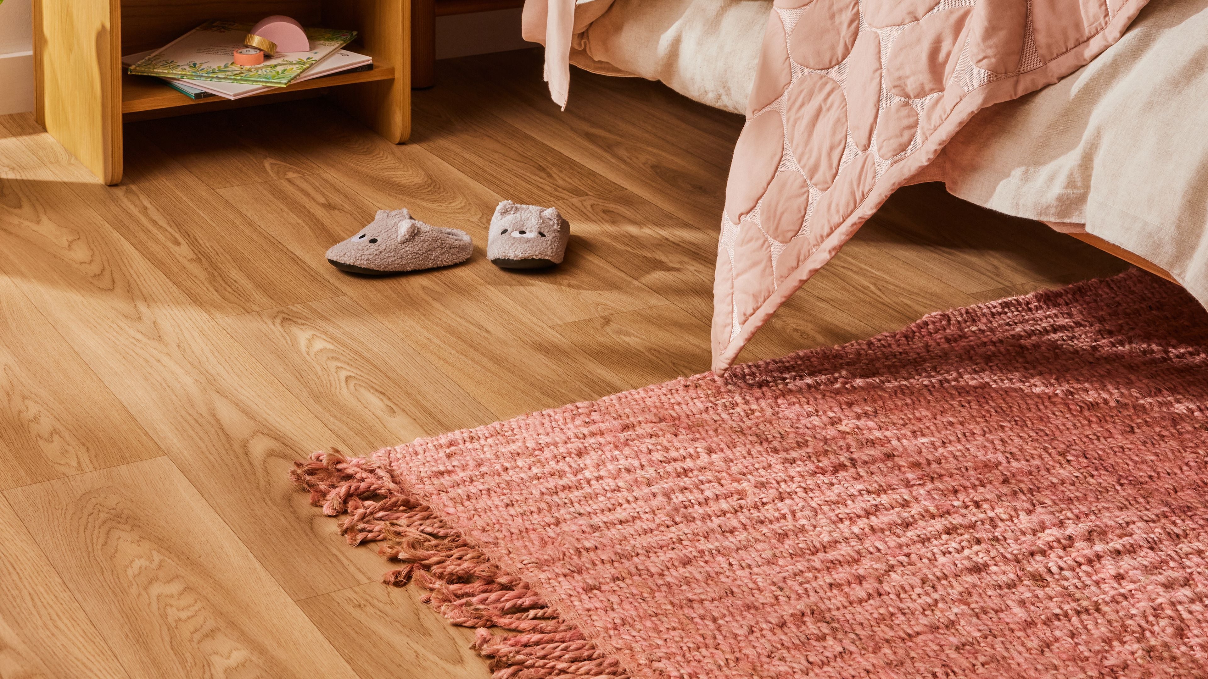 Bedroom with light wood flooring, pink rug, pink quilt, two grey slippers, and a wooden nightstand with books. Cozy and inviting decor.