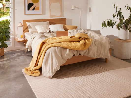 Bedroom with a wooden bed, light grey bedding, mustard throw, and beige rug on concrete floor. Sunlit room with plants and framed art.