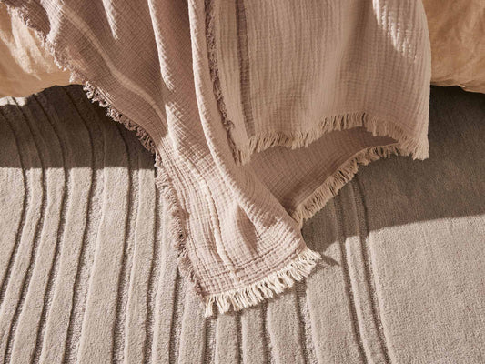 Beige textured throw with fringed edges draped over a soft, light grey rug with striped patterns, illuminated by warm natural light.