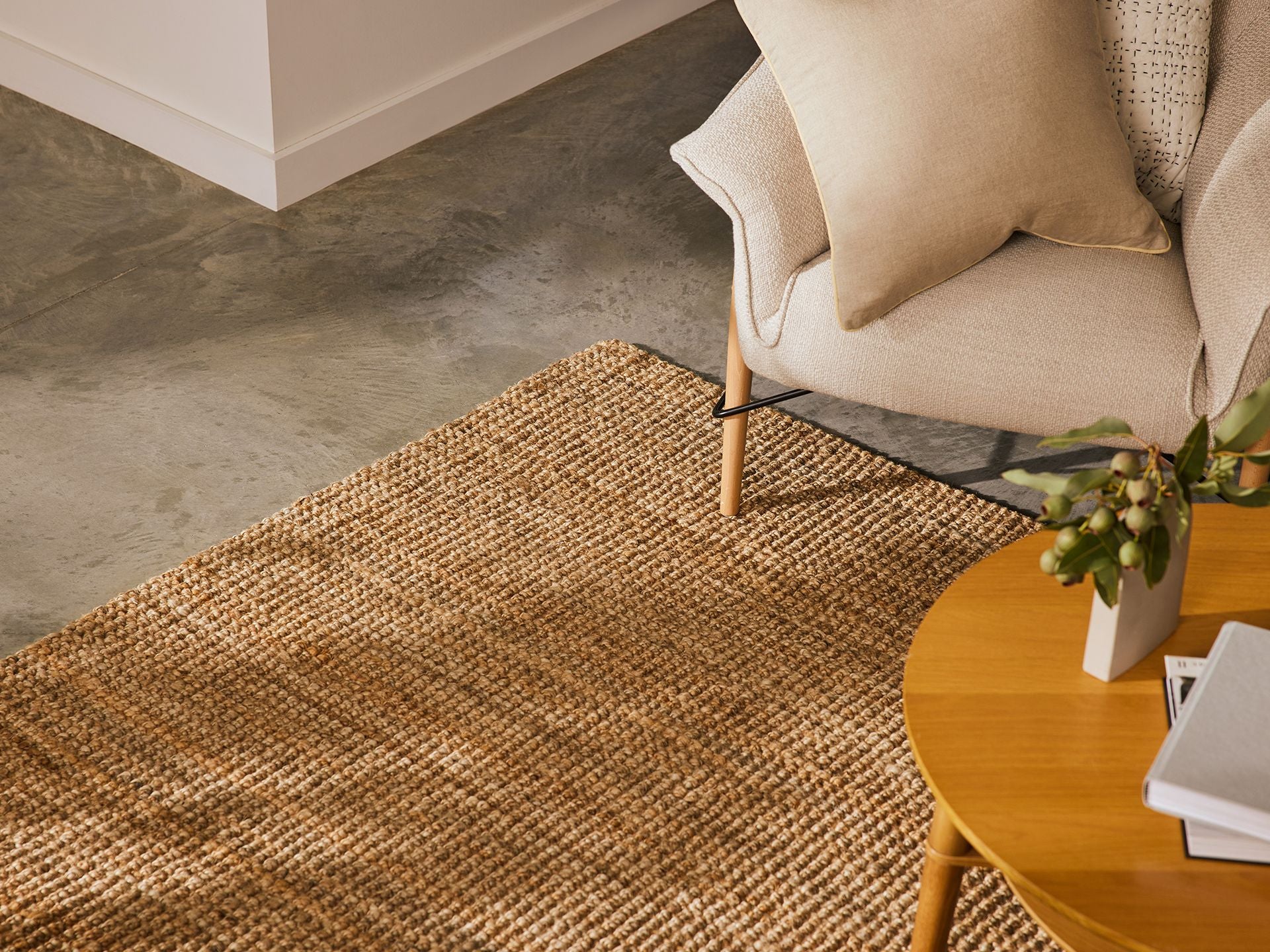 Cosy living room with a beige armchair, woven rug, and round wooden table on concrete floor. Neutral decor and books create a relaxing ambience.