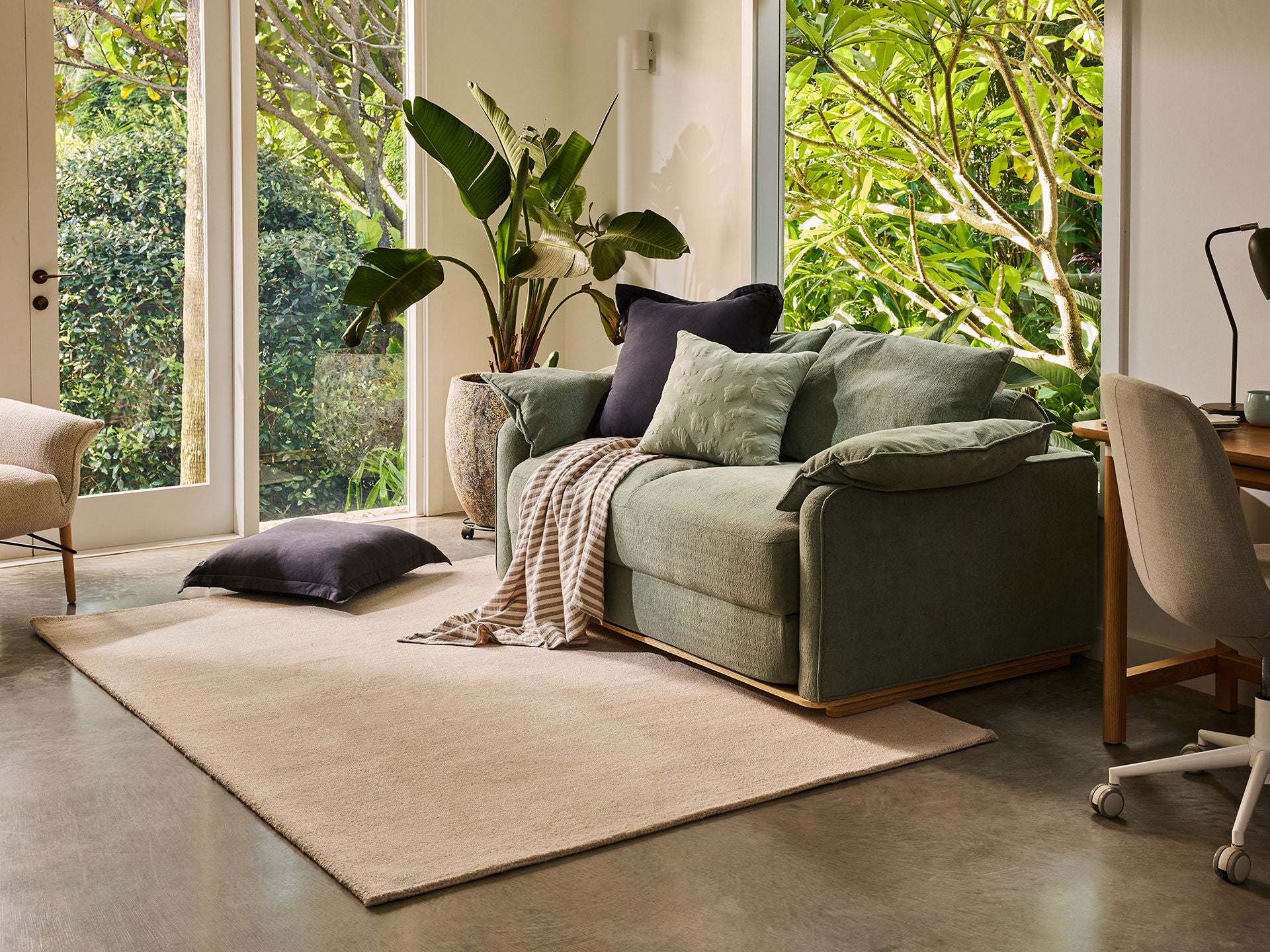 Modern living room with a green sofa, beige rug, and a light grey chair at a desk. Large plants and floor-to-ceiling windows add greenery.