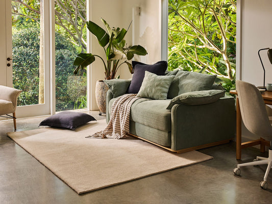 Modern living room with a green sofa, beige rug, and a light grey chair at a desk. Large plants and floor-to-ceiling windows add greenery.