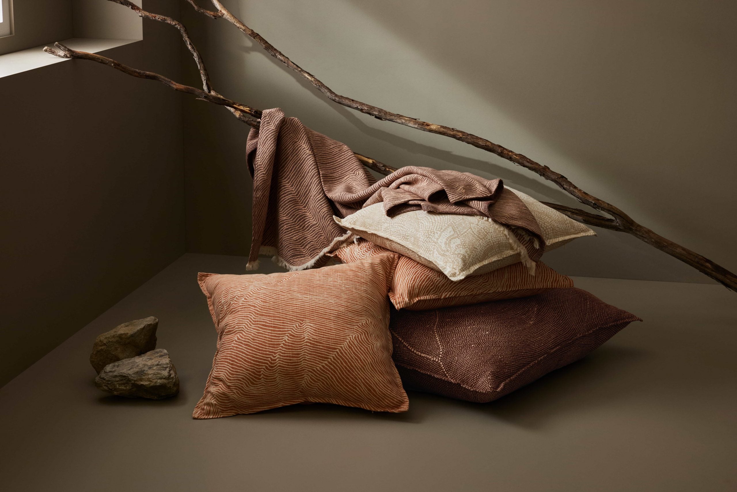 Pillows and throws in earthy tones stacked on a neutral floor, with rustic sticks and stones nearby. Natural, cosy home decor aesthetic.