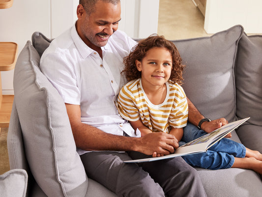 Man and child reading on a grey couch. The man wears a white shirt, and the child wears a yellow striped shirt. Cozy family scene.