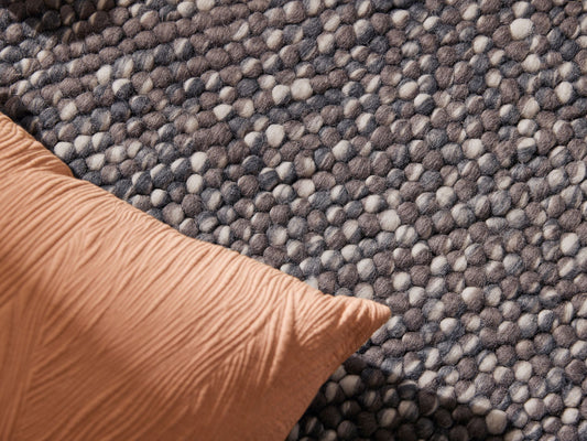 Close-up of a textured woven rug with a mix of grey and beige tones, and an orange cushion on one corner. Perfect for cosy home decor.
