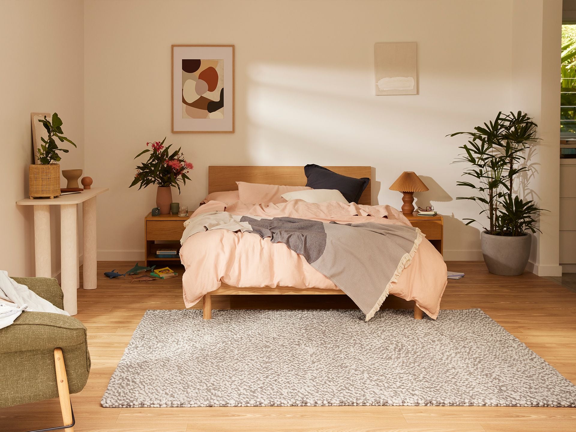 Bedroom with a wooden bed, peach and grey bedding, two bedside tables, plants, abstract art, and a textured grey rug on wooden flooring.