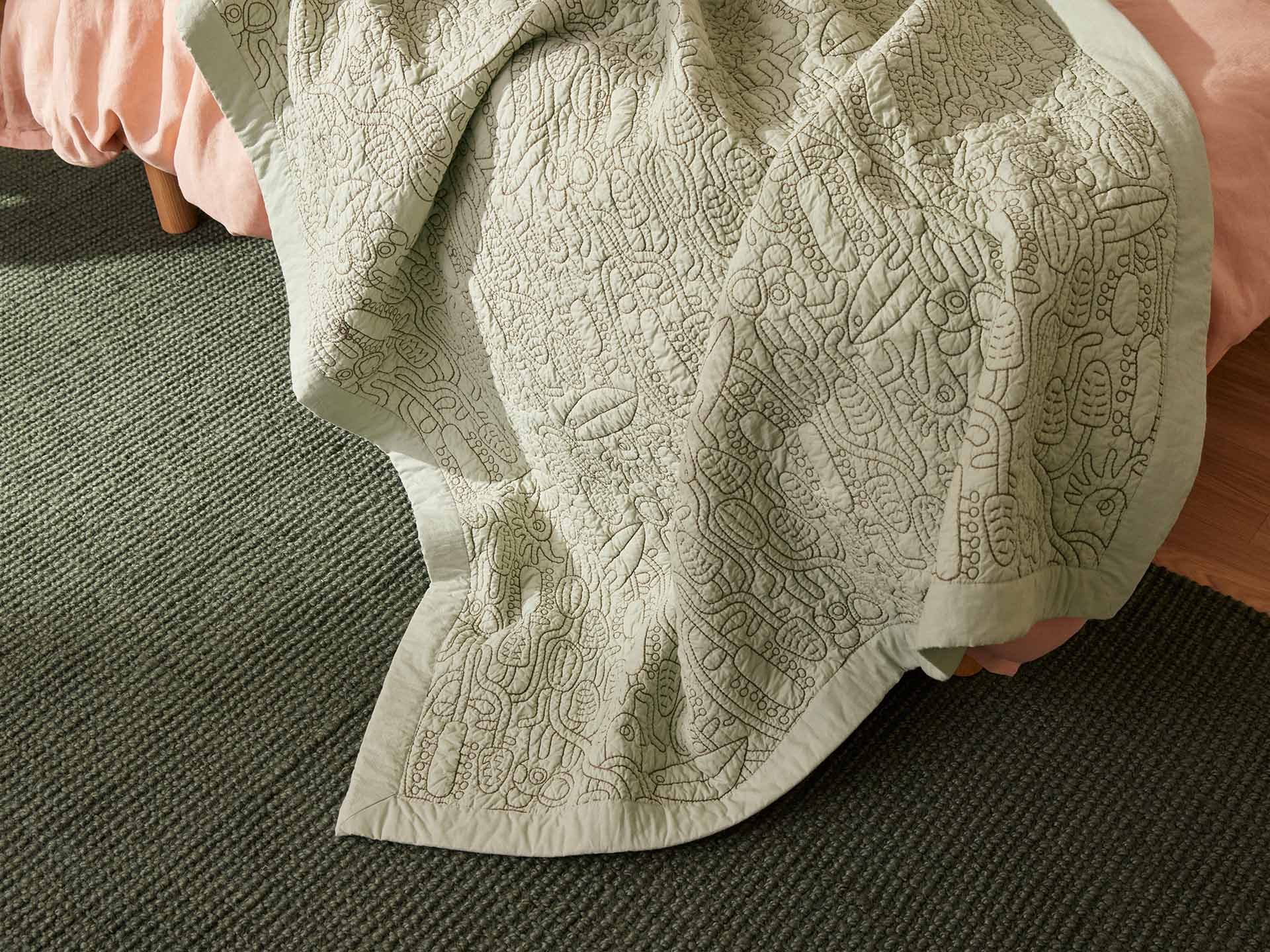 Sage green quilt with intricate stitching draped over a bed, against a dark green carpet.