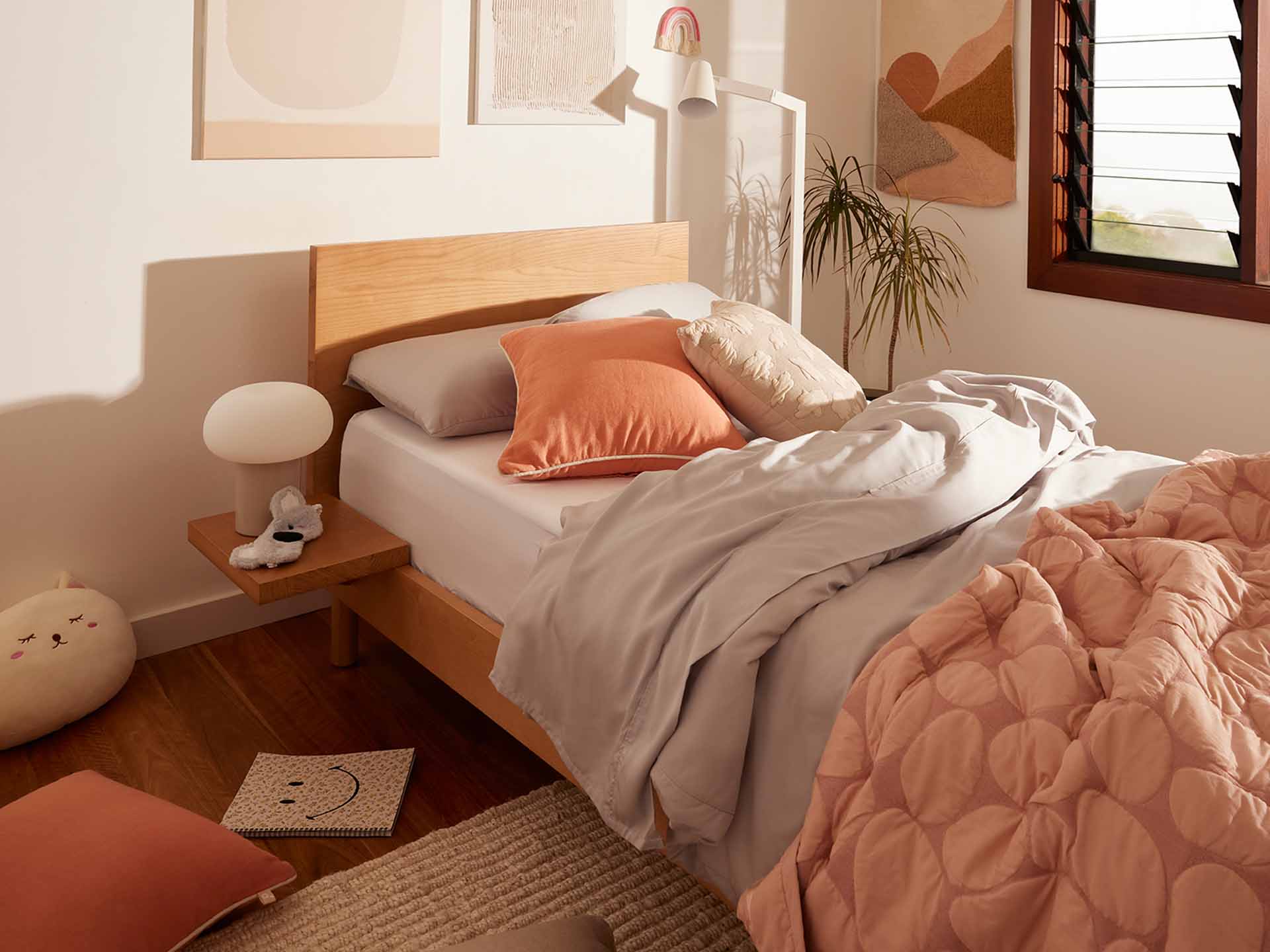 Cosy bedroom with a wooden bed, grey and peach bedding, a small side table with a lamp, and wall art. Light streams through wooden shutters.