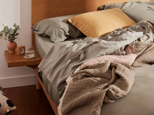 Unmade bed with light grey sheets, beige blankets, and a mustard pillow on a wooden frame. A small shelf holds a plant, clock, and glass of water.