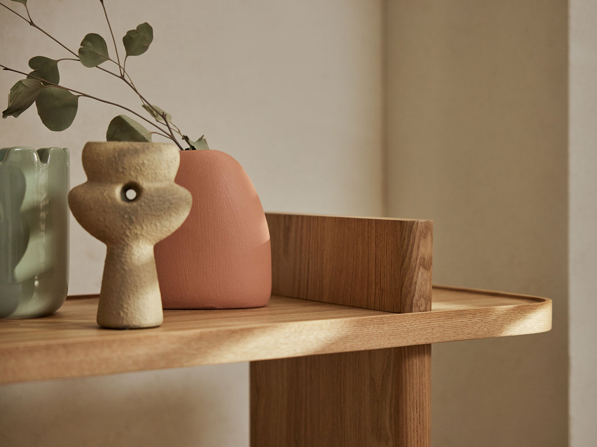 Wooden shelf with a beige abstract vase, coral vase, and green vase with leaves, set against a light wall.