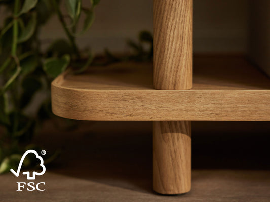 Close-up of a light brown wooden shelf edge, with a round post, surrounded by green leaves. FSC logo in the corner.
