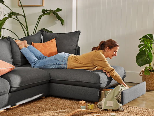 Woman relaxes on a dark grey sectional sofa with orange cushions, exploring a storage drawer. Indoor plants and a woven rug enhance cosiness.