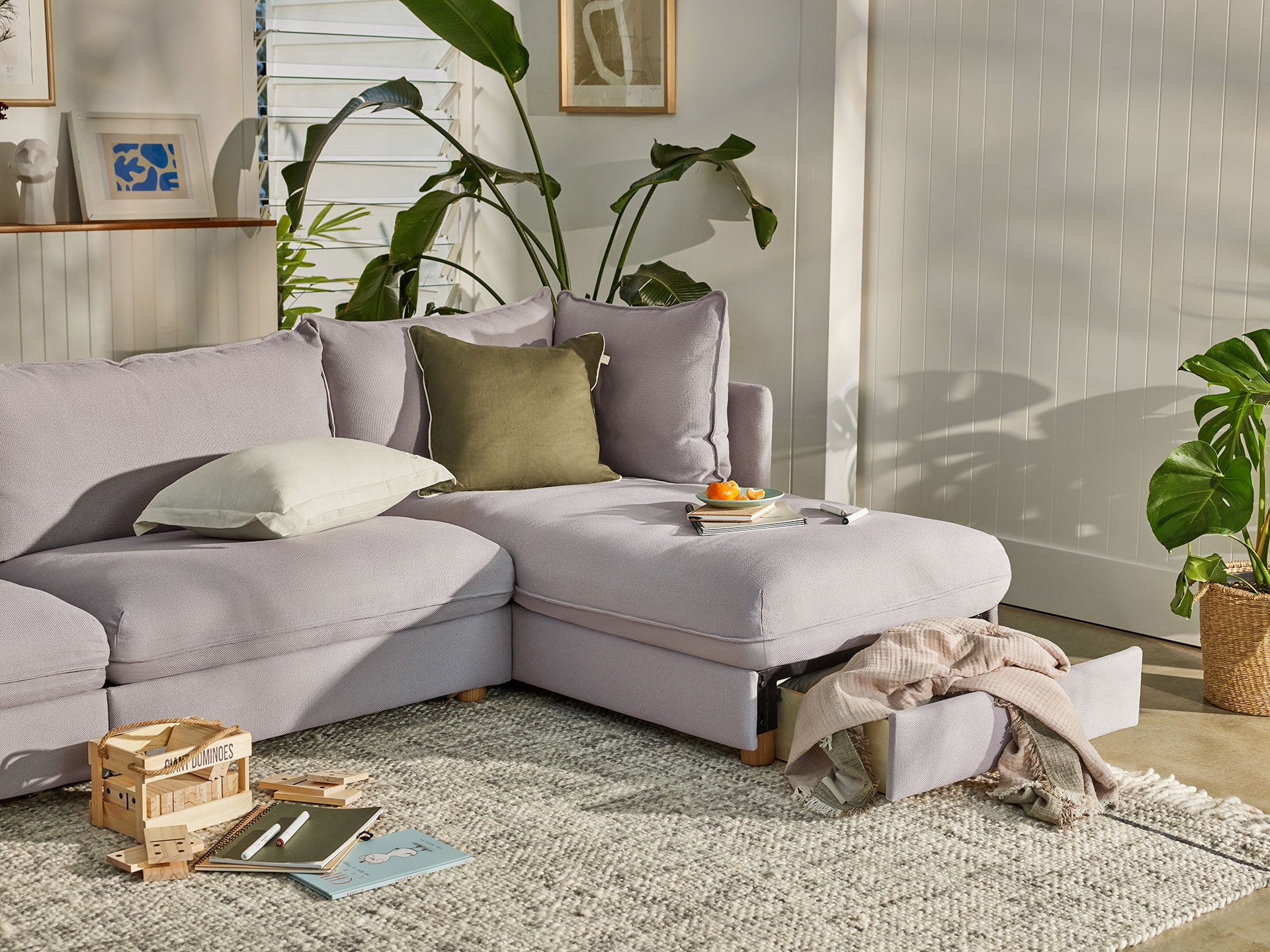 Light grey sectional sofa with storage chaise, olive and white cushions, surrounded by indoor plants, a textured rug, and muted decor elements.