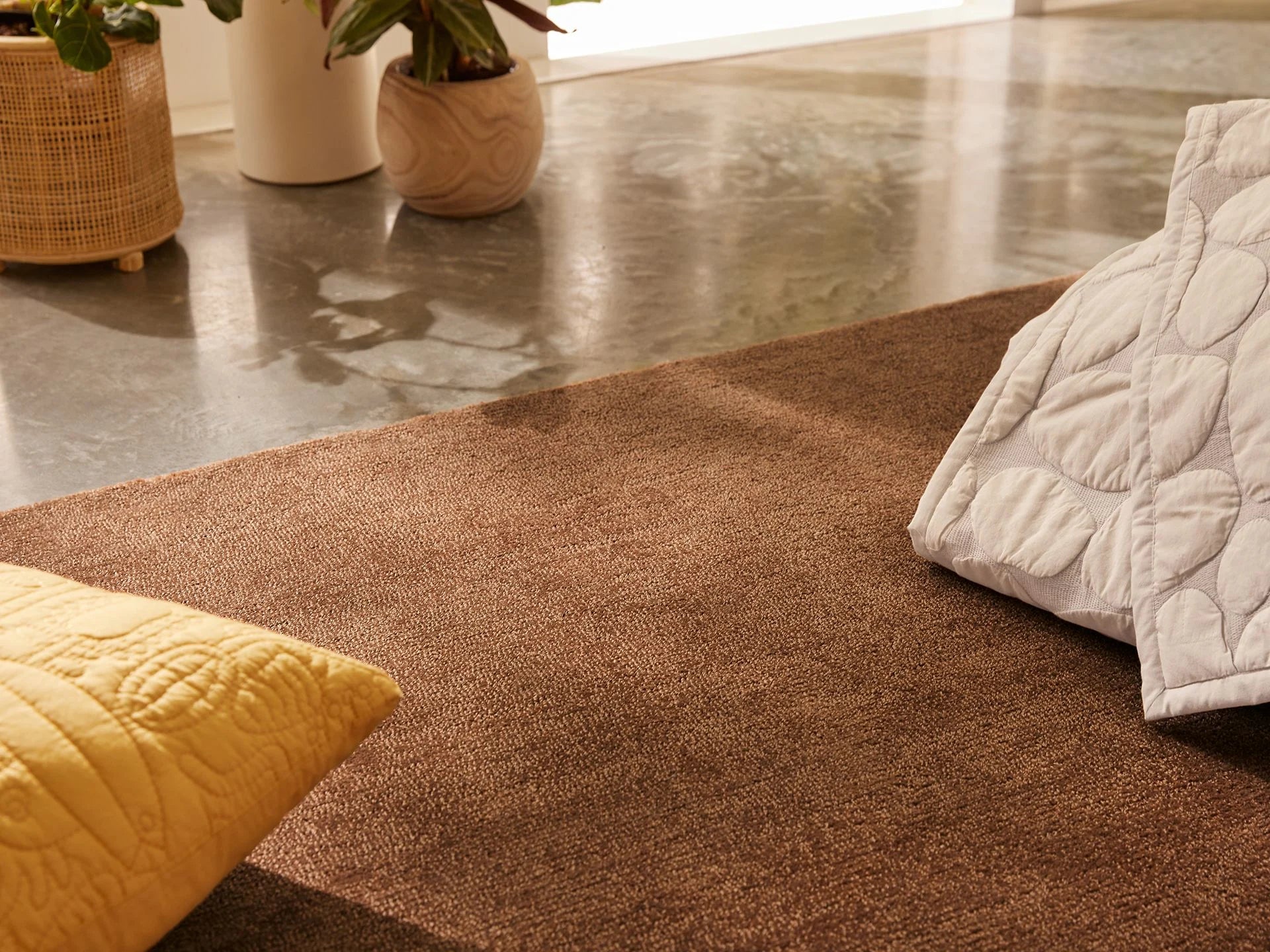Brown rug on polished concrete floor with quilted pillows, one yellow and one white. Potted plants in woven and ceramic pots nearby.