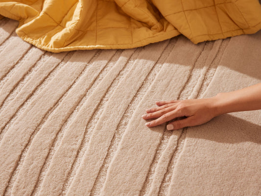 Hand touching a textured light beige carpet with raised stripes, next to a crumpled yellow blanket. Warm, cosy interior setting.