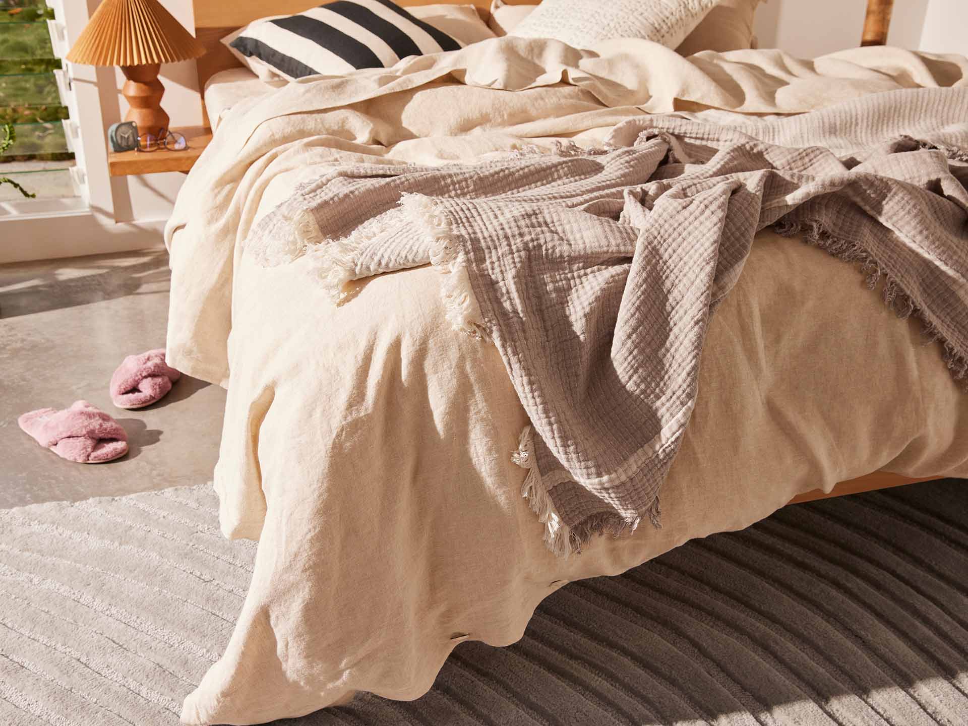 Cosy bedroom with a cream quilt, grey throw, and striped pillows on a wooden bed. Pink slippers on a grey rug beside a small table lamp.