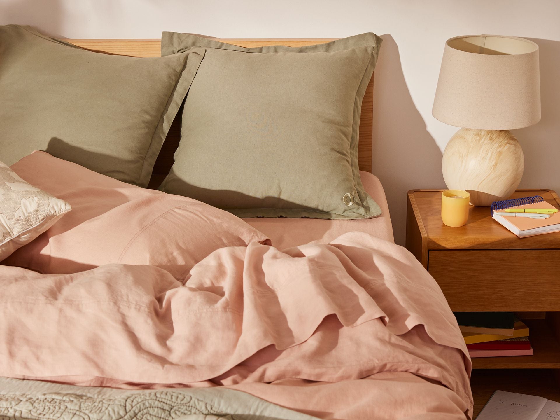 Bedroom scene with a made wooden bed featuring olive green pillows and a pink quilt. A bedside table holds a beige lamp, a yellow cup, and books.
