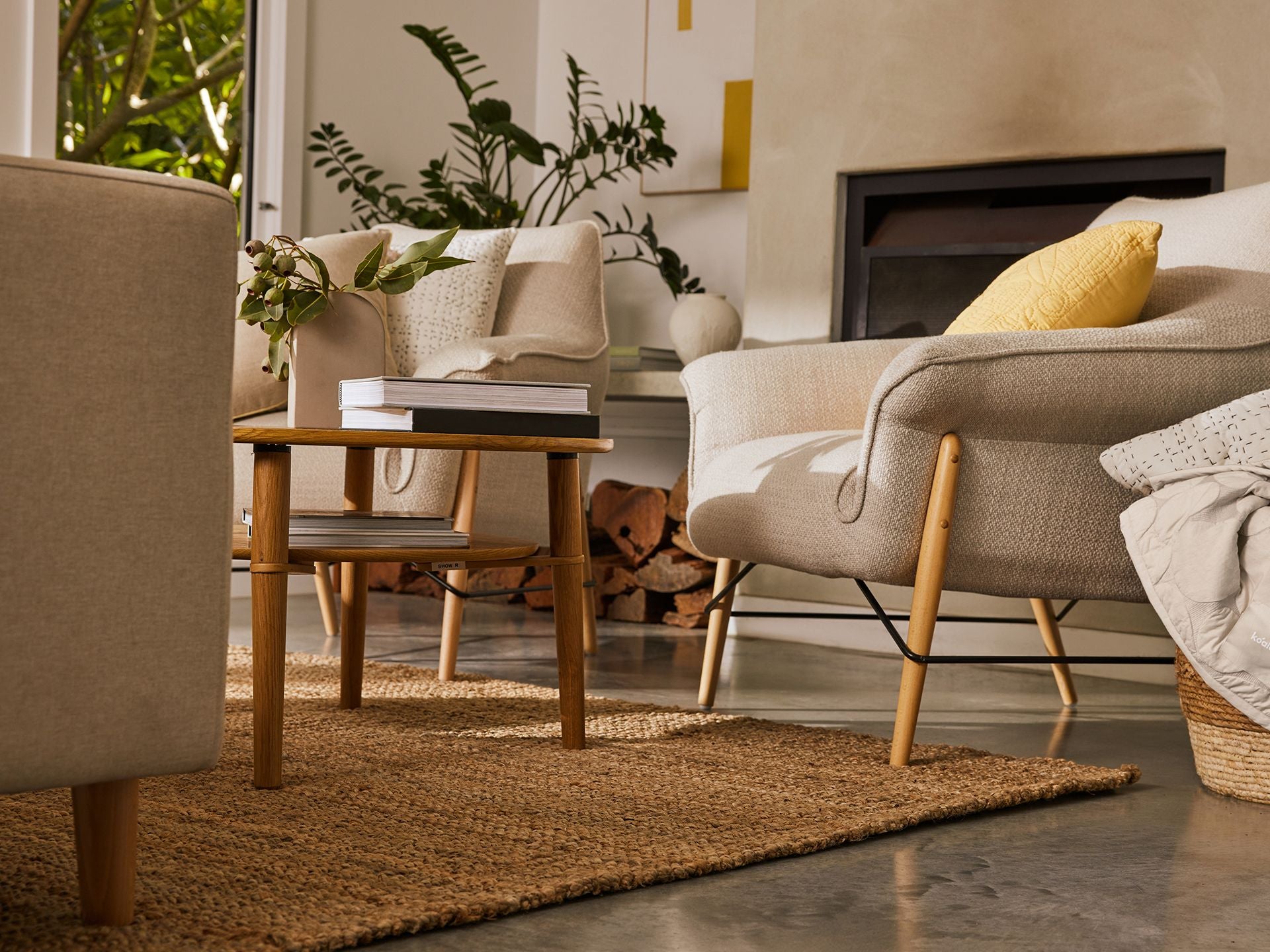 Cosy living room with beige armchairs, a woven rug, wood coffee table, and fireplace. Decor includes plants, books, and decorative cushions.