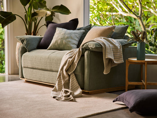 Sage green couch with assorted cushions and a draped blanket. Wooden side table with vase; indoor plants by large window, neutral carpet.