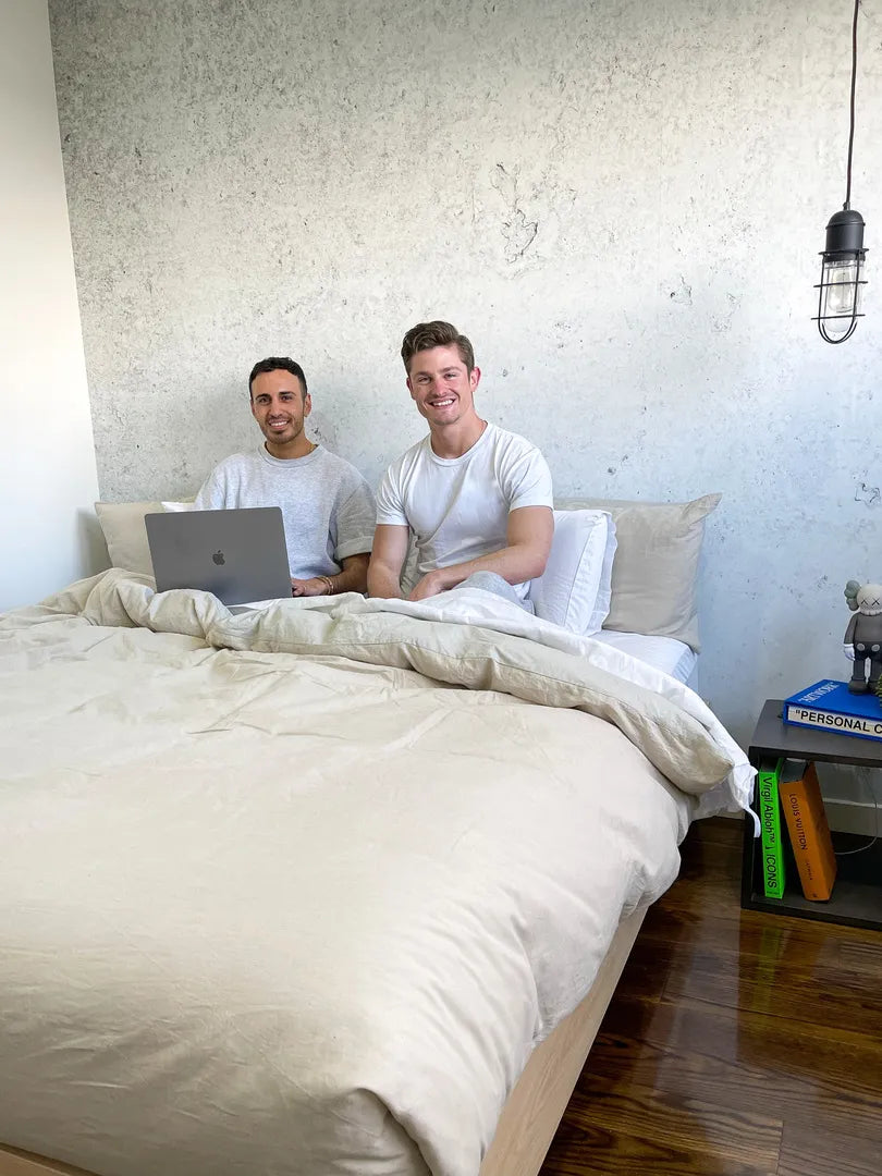 Two men sitting in bed with beige bedding, one using a laptop. The room has a textured grey wall, wooden floor, and bedside table with books.