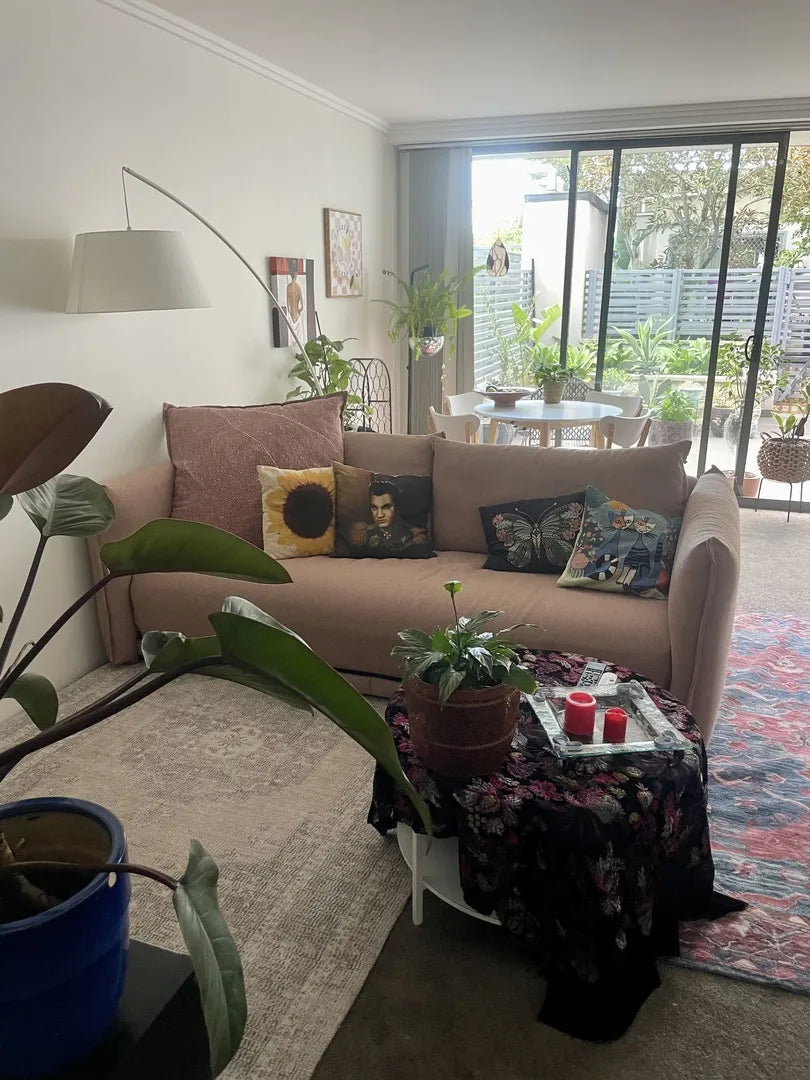 Living room with a beige couch and colourful cushions, glass-topped table with plants, dining area in the background, and sliding glass doors.