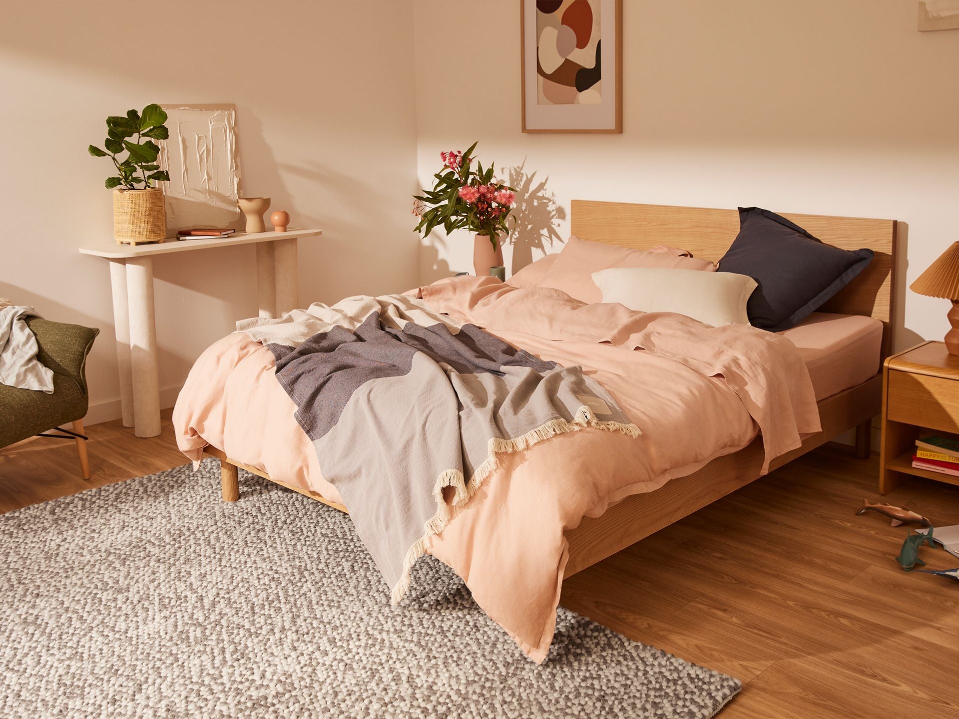 Modern bedroom with a wooden bed, pink and grey bedding, green chair, and light grey rug on wooden floor. Decor includes a plant and artwork.