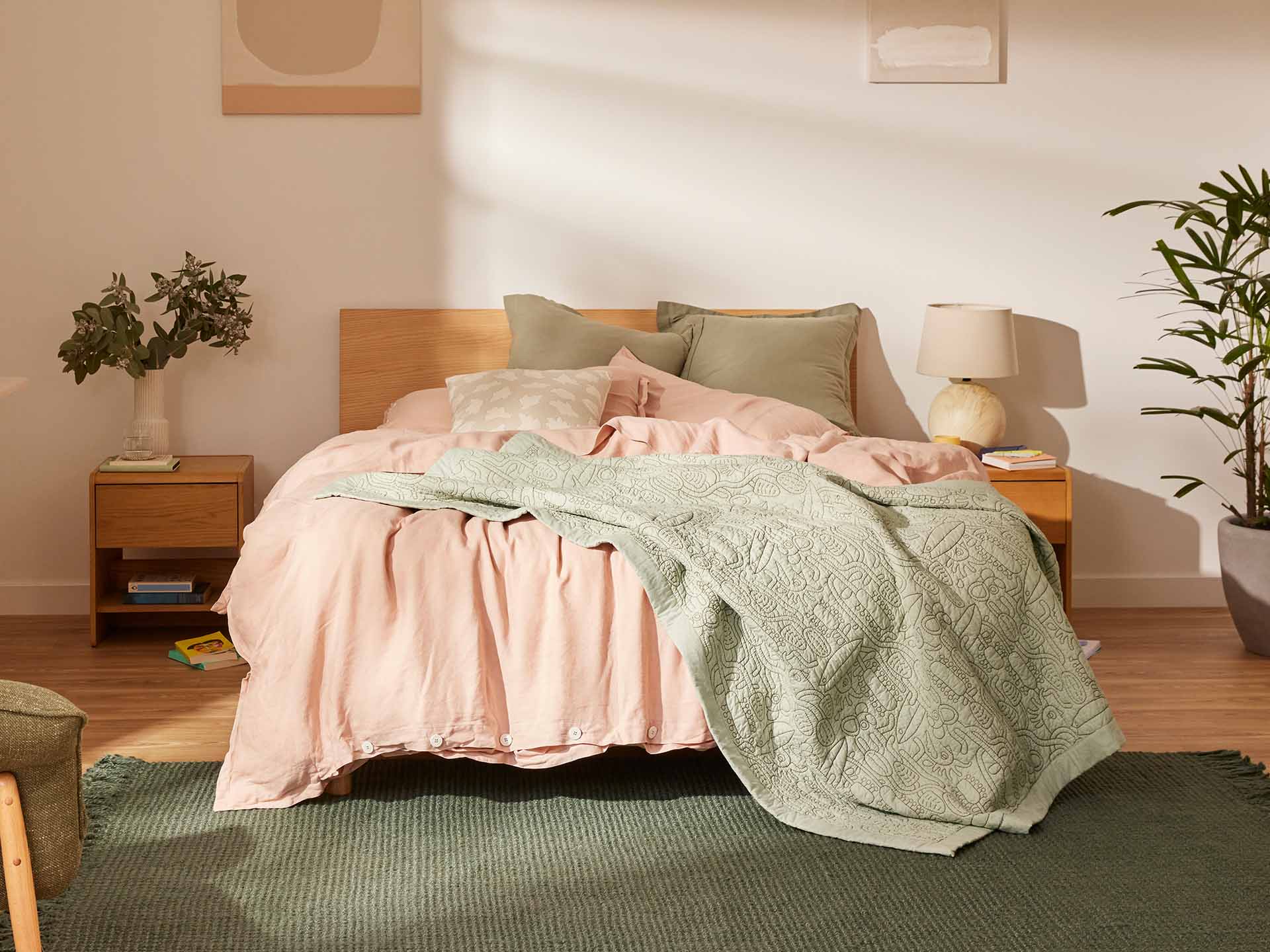 Bedroom with a wooden bed, pink bedding, green quilt, bedside tables, white lamp, and plant. Light-filled room with paintings on the wall.